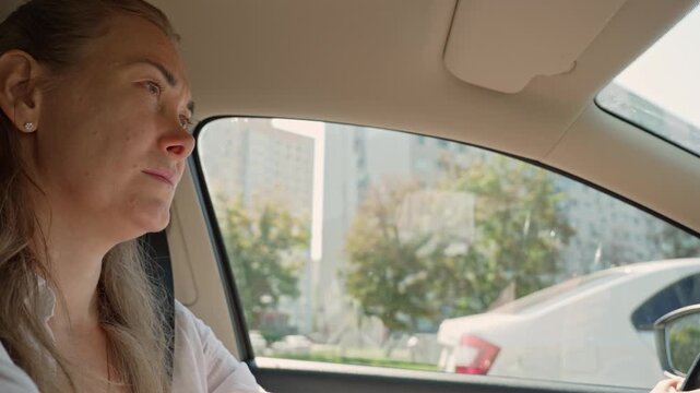 Middle-aged woman with a pensive and tired expression driving her car through a sunlit city, looking out the window while sitting in traffic, conveying a moment of reflection and weariness