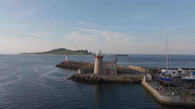 Stone lighthouse stands on the breakwater as the drone moves across the harbour entrance toward distant Ireland&rsquo;s Eye island.