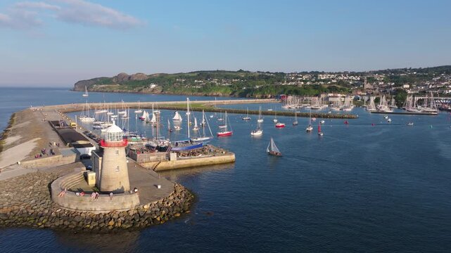 Visitors walk around the Howth harbour breakwater lighthouse as sail craft navigate the sheltered entrance on a clear summer afternoon.