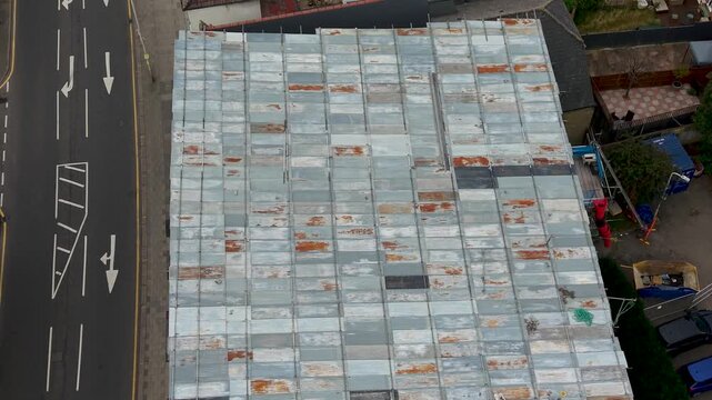 Drone flyover of UK low-rise flats showing corrugated sheet metal roof repairs beside a town road as cars pass on left-hand traffic. Top-down urban housing, maintenance, infrastructure.