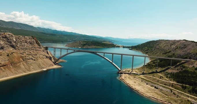 Krk Bridge crossing turquoise Adriatic water between rocky coastal hills in Croatia. Mountains, islands, shoreline, and blue summer sky fill the distance.