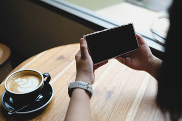 Naklejka premium An over the shoulder view of a person holding a smartphone horizontally with a blank black mockup screen at a wooden cafe table next to a cup of coffee.