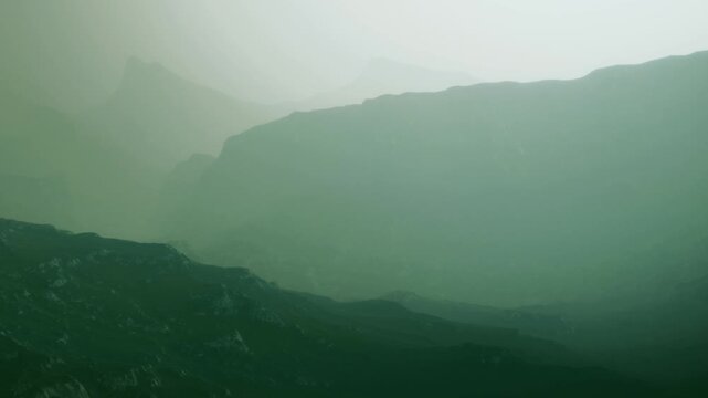 Mountains appear shrouded in mist during dusk. The landscape shows different shades of green. The scene captures the fading light and outlines of the mountains in the background.