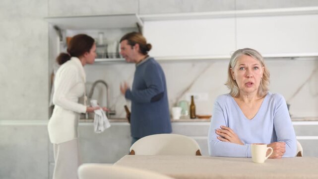 Upset old woman sitting in kitchen and man and woman arguing in background 