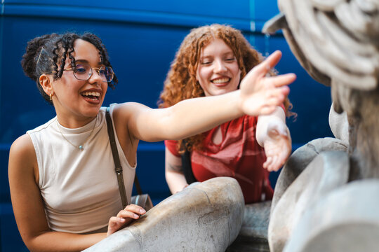 Two happy young female friends, one with curly dark hair and glasses and the other with ginger hair, laugh as they reach out to touch a historic stone statue.