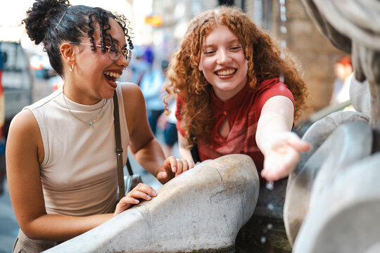 Two happy young female friends laugh together on a city street. One friend reaches out to touch the cool, refreshing water flowing from an old stone fountain.
