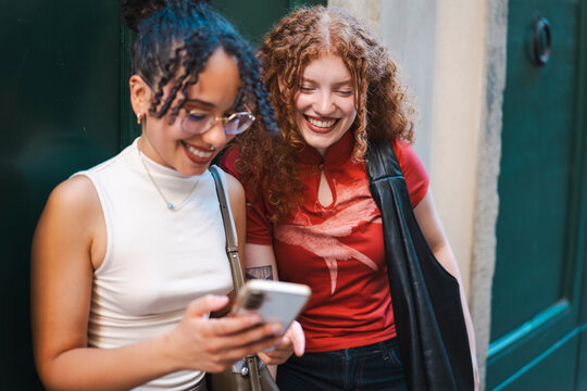 Two cheerful young female friends, one with curly red hair and the other wearing glasses, laugh together while looking at a smartphone screen on a city street.