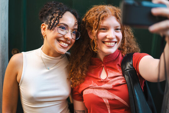 Two cheerful young women with beautiful curly hair smile warmly as they take a selfie together. The diverse friends pose close to each other in front of a green wall.