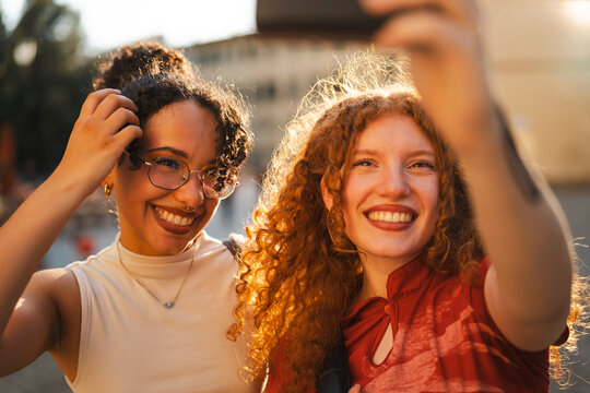 Two diverse young female friends with curly hair smile happily while taking a selfie together with a smartphone outdoors during a beautiful golden hour sunset.