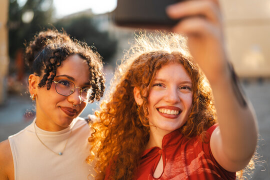 Two happy diverse young women with curly hair smile warmly while taking a selfie together on a smartphone outdoors in the beautiful golden hour sunlight.