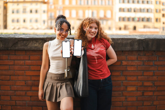 Two smiling young women stand outdoors by a brick wall and hold up two smartphones with blank, white screens. Perfect for app mockups and mobile interface designs.