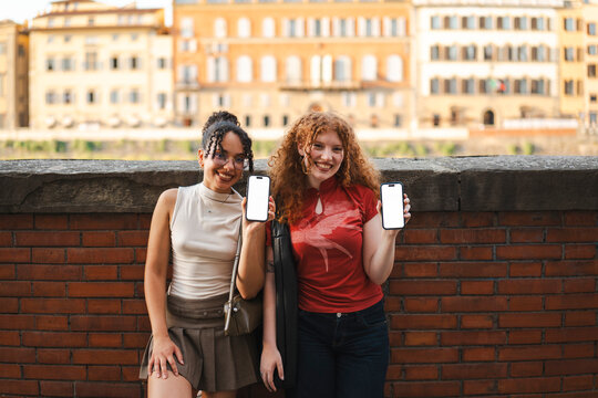Two smiling diverse young women show their blank smartphone screens while standing against a brick wall in a historic European city. Ideal for mobile app mockup.