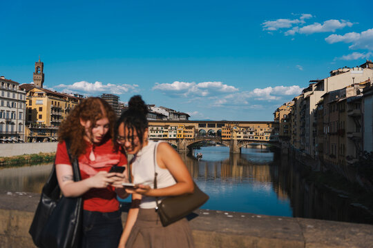 Two young female friends stand out of focus in the foreground, looking at their smartphones with a famous historic bridge over a river in the background.