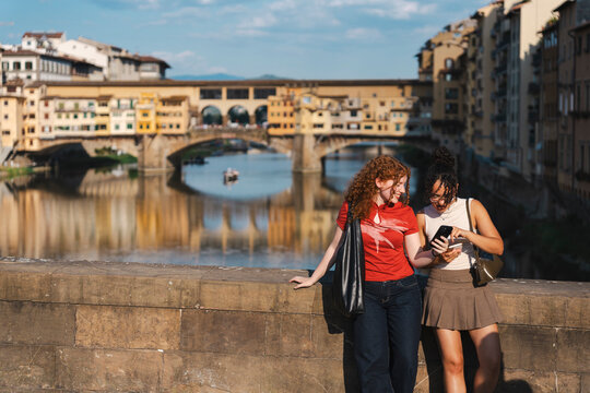 Two diverse young women smile and look at a smartphone while standing on a stone bridge in Florence, Italy, with a historic bridge over a river behind them.