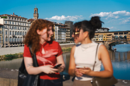 Two young female tourists holding smartphones talk in the blurred foreground, with the historic city of Florence, Italy and its river bridge in sharp focus.