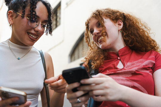 Two cheerful young female friends with curly hair stand outdoors, smiling and laughing as they look at a smartphone screen together on a charming city street.