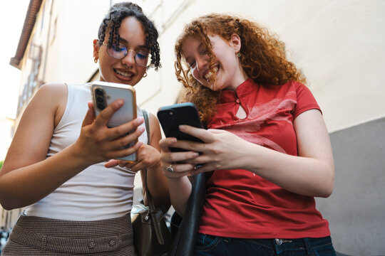 Two cheerful diverse female friends stand together on a city street, smiling and laughing as they look at their smartphones and share entertaining online content.