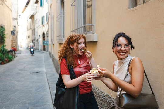 Two cheerful young female friends laugh and eat Italian gelato together on a narrow historic city street, sharing a happy moment during their summer vacation.