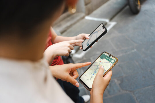 Two friends standing on an urban cobblestone street use map navigation apps on their smartphones to find directions. They point at the screens to plan their route.