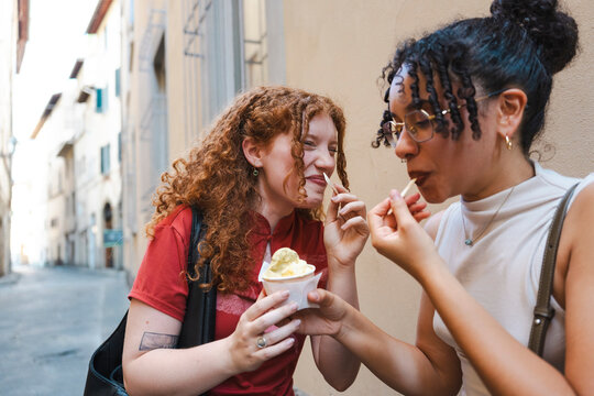 Two diverse young female friends enjoy eating cold Italian gelato together on a narrow European street. They are smiling, laughing, and sharing a sweet dessert.