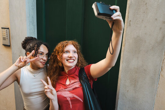 Two smiling diverse young female friends pose and make peace signs while taking a selfie together with a vintage digital camera in a narrow city street.