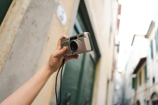 A tourist holds a vintage silver compact film camera in their hand, pointing it upwards to take a photo in a narrow old European street during a summer vacation.