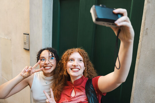 Two happy young female tourists smile and make peace signs while taking a selfie together on a compact digital camera in front of a rustic green wooden door.
