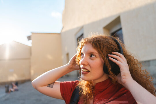 A cheerful young redhead woman wearing headphones listens to music outdoors during a bright sunset on a city street, looking away with a joyful expression.