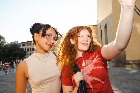 Two cheerful diverse female friends pose and make playful faces while taking a selfie together in a sunny, historic European city square during summer vacation.