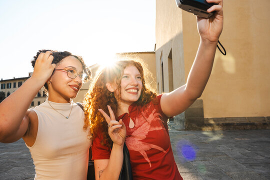 Two happy diverse young female friends smile and pose for a selfie with a camera. They stand on a sunny historic street during golden hour on their vacation.