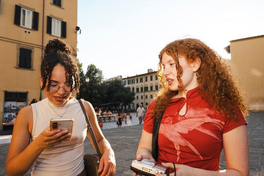 Two young female tourists explore a historic European city square during sunset. One looks at her smartphone while her red-haired friend holds a camera and talks.