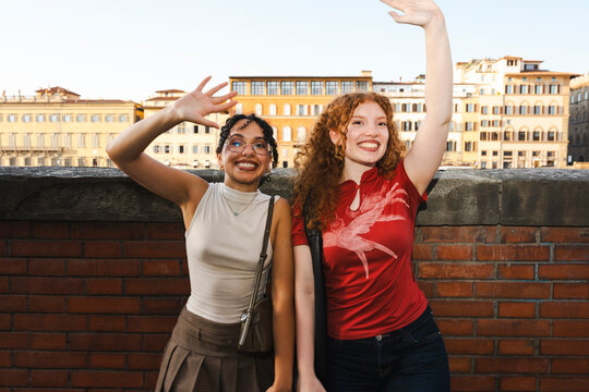 Two cheerful young diverse women tourists smiling and waving at the camera while standing by a brick wall with historic European buildings in the background.
