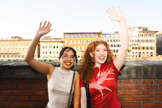 Two happy diverse young female friends wave their hands and smile warmly while posing together on a vacation in a historic European city during sunset.