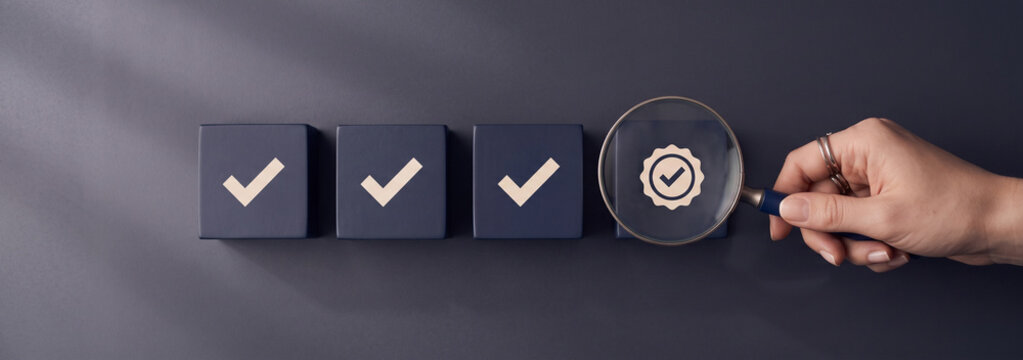 Hand holding magnifying glass over check mark cubes. Quality assurance and business compliance concept.