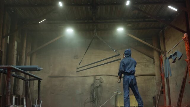 Industrial worker spray painting metal rods with protective coverall and respirator while suspended parts hang under overhead hoist in high-ceiling warehouse, with worker, spray, painting