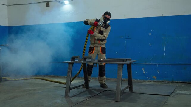 Technician lifting yellow black striped hose behind metal workbench inside smoke filled industrial workshop with protective coverall respirator goggles and gloves
