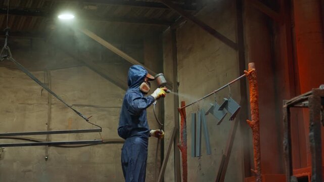 Industrial worker spray painting suspended metal parts inside dim warehouse, wearing protective coverall, respirator, gloves, and hood while coating brackets with orange red mist
