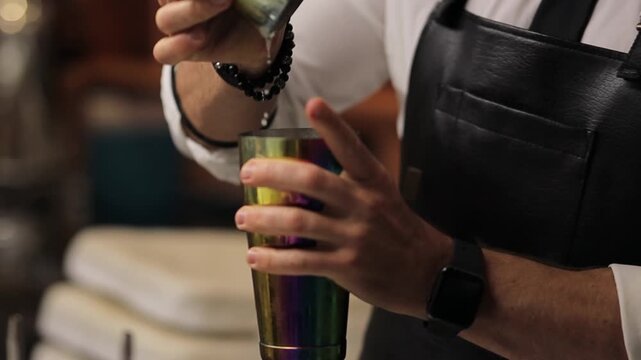 A bartender in a white shirt and black apron holds a metal cocktail shaker in his hands.