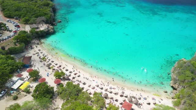 High overhead aerial view of Grote Knip Beach Curacao reveals full crescent turquoise bay packed with swimmers sun umbrellas and lush green cliffs