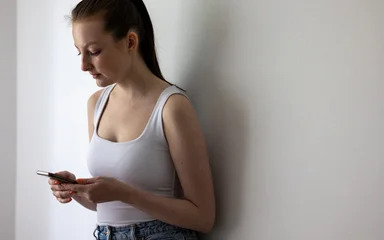 Young Woman Leaning Against Wall While Looking at Smartphone, Concept of Digital Stress and Social Media Anxiety © lightpoet