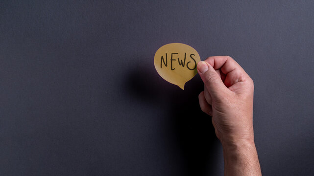 Hand holding a speech bubble with the word news written in black on a dark background at an indoor location during the day