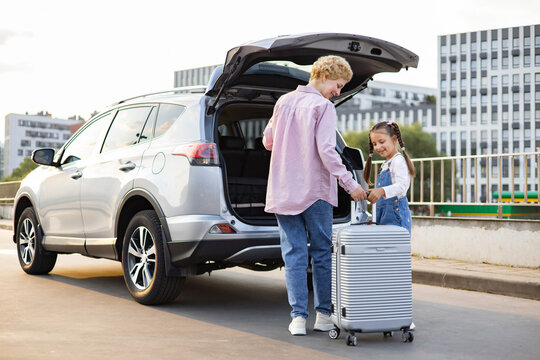A Caucasian mother and her daughter are loading a suitcase into the back of a silver SUV parked on a city street