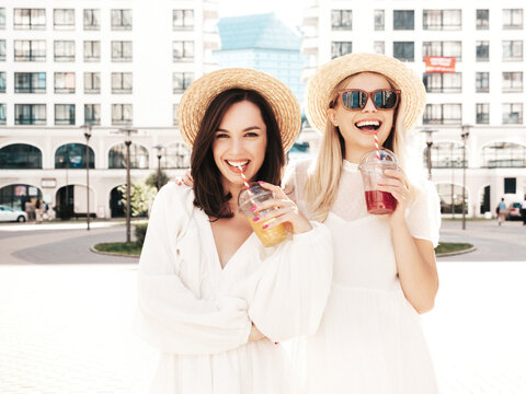Two young beautiful smiling hipster female in trendy summer white dress and straw hats. Sexy carefree women holding and drinking fresh vegetable cocktail smoothie drink in plastic cup with straw
