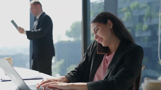 Cheerful businesswoman in office using laptop and phone typing with professional expression, businessman by window, workplace communication technology corporate multitasking suit