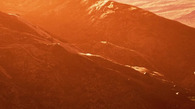 Mountains are visible in a barren area during sunset. The sky reflects a bright orange hue casting shadows on the uneven terrain. The scene captures a moment of daily change in nature.