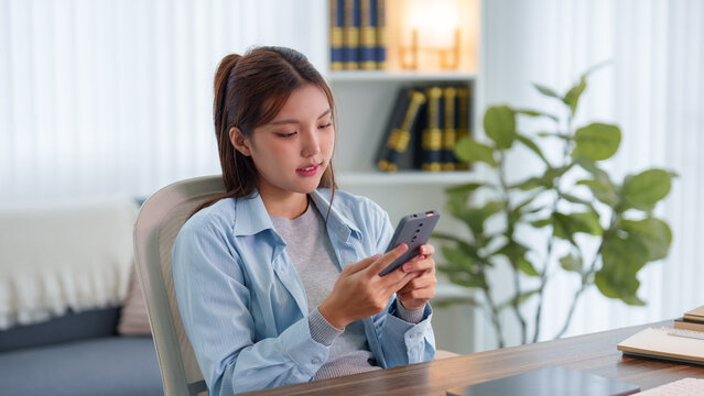 Young Asian woman using smartphone for mobile learning while studying at home desk with notebook