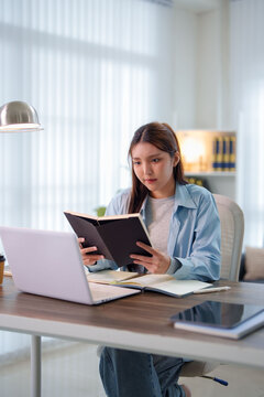 Young Asian student reading notebook while studying with laptop at home desk in bright interior