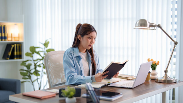 Young Asian woman reading notebook while studying with laptop at modern home desk with copy space