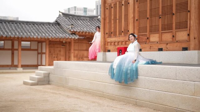 Traditional Korean architecture frames serene figures. One woman sits gracefully, draped in flowing blue-white attire. Another stands nearby, wearing soft pink and white