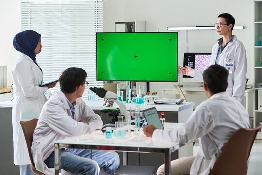 Wide shot of diverse scientists discussing research results in laboratory meeting, presenting on green screen monitor. Useful for healthcare innovation, biotech training, medical marketing